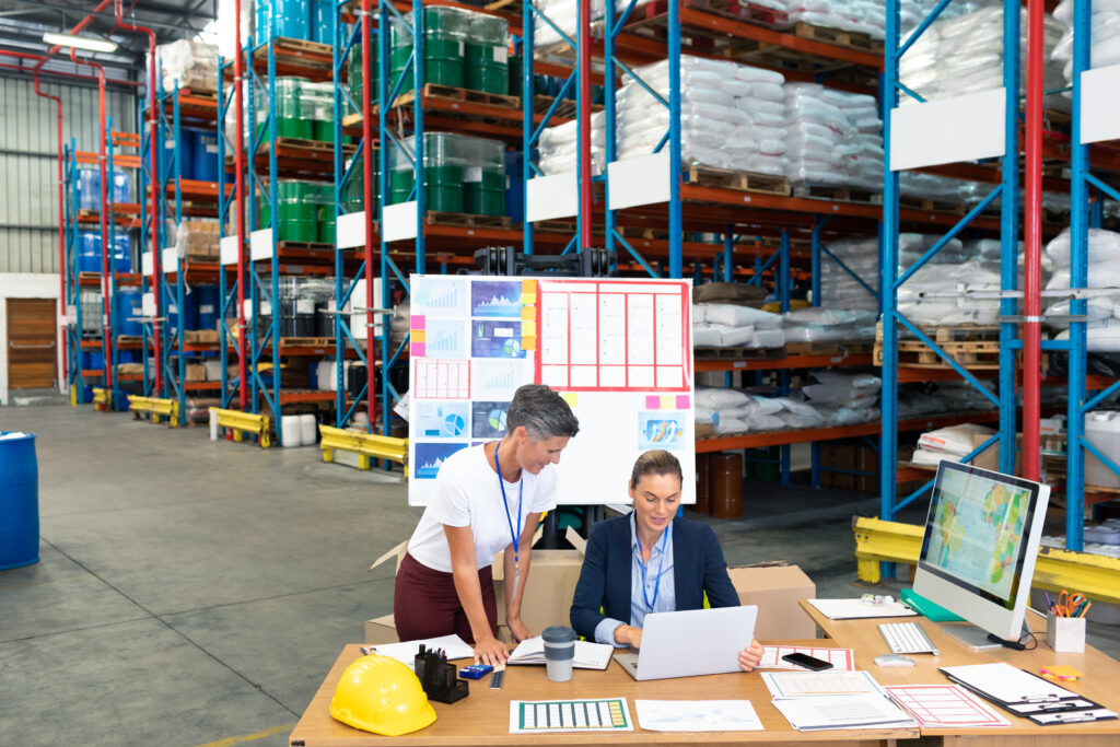 Supply Chain Management Logistics That Scale With Your Business high angle view of beautiful caucasian female manager with her coworker discussing over laptop at desk in warehouse. this is a freight transportation and distribution warehouse. industrial and industrial workers concept