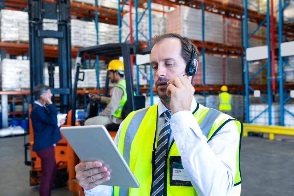 close up of handsome caucasian male supervisor using digital tablet while talking on headset in warehouse. diverse colleagues communicating in the background. this is a freight transportation and distribution warehouse. industrial and industrial workers concept