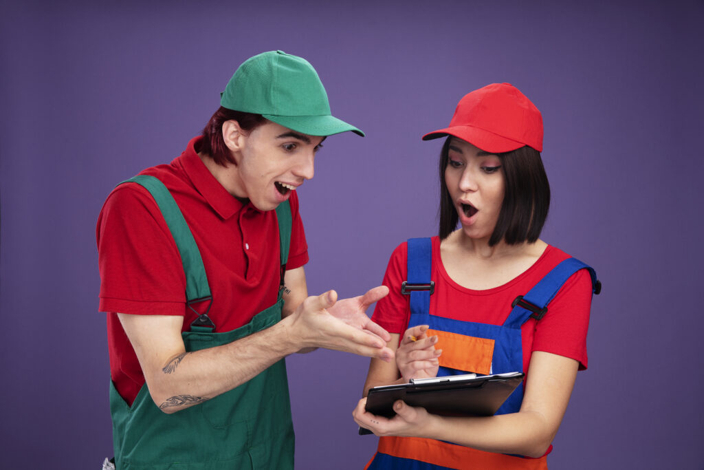young couple in construction worker uniform and cap surprised girl holding pencil and clipboard excited guy pointing with hands at clipboard both looking at clipboard isolated on purple background