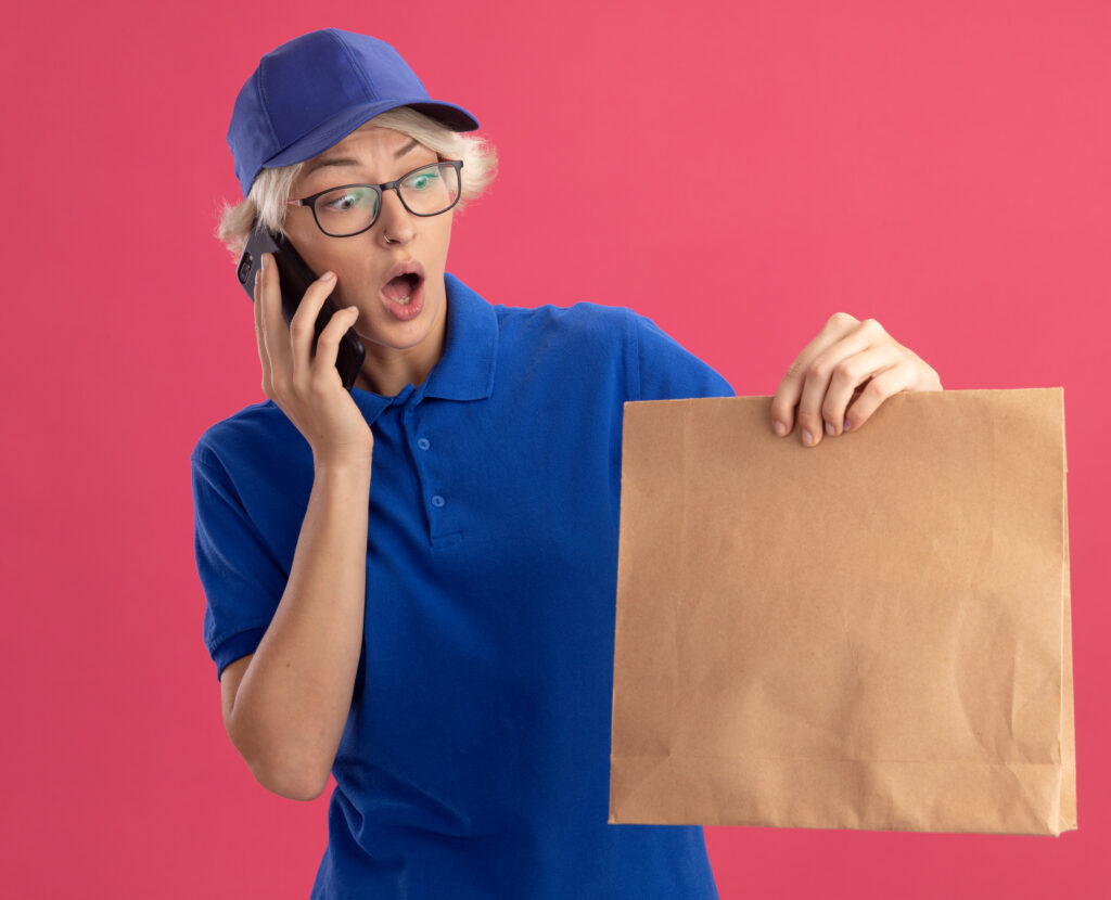 What Makes Customers Complain About Delivery young delivery woman in blue uniform and cap holding paper package looking surprised and amazed while talking on mobile phone over pink background